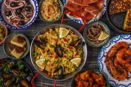 Typical spanish tapas concept. Concept include slices jamon, bowls with olives,  anchovies, spicy potatoes, mashed chickpeas, shrimp, calamari, manchego with quince marmalade, pans with tortilla, paella, mussels  on a wooden table.の写真素材