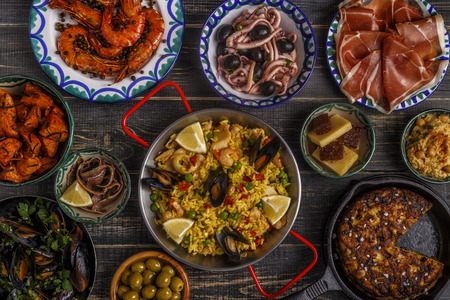 Typical spanish tapas concept. Concept include slices jamon, bowls with olives,  anchovies, spicy potatoes, mashed chickpeas, shrimp, calamari, manchego with quince marmalade, pans with tortilla, paella, mussels  on a wooden table.の写真素材