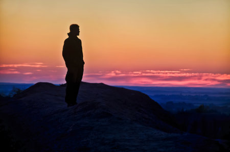 Silhouette of a man standing on top of a mountain and looking at the sunsetの写真素材