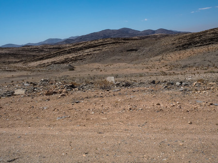 Road trip through rough dusty unpaved road with car tyre track through dried desert climate and hard rock mountain landscape background with clear blue sky, Namibiaの写真素材