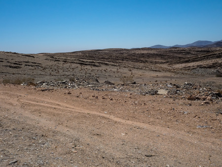 Road trip through rough dusty unpaved road with vehicle tyre track through drought desert climate and hard rock mountain landscape background with clear blue sky and dried plant, Namibiaの写真素材