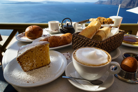 Beautiful breakfast with Aegean sea view and morning sunlight including cappuccino cup, cake, baguette, croissant, boiled egg, hot tea and chocolate brioche bun on white marble table, Oia, Santorini, Greeceの写真素材
