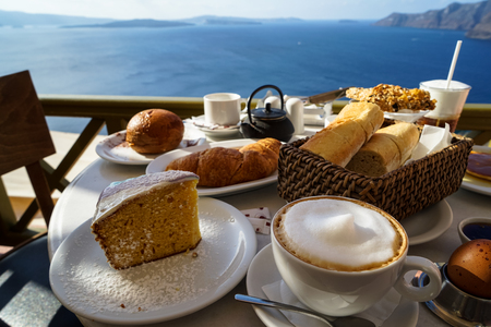 Enjoy island breakfast with beautiful Aegean sea view and morning sunlight including cappuccino cup, cake, baguette, croissant, boiled egg, hot tea and chocolate brioche bun on white marble table, Oia, Santorini, Greeceの写真素材