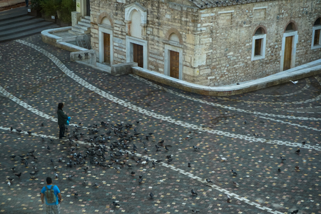 A man feeding pigeons at beautiful floor pattern Monastiraki square in front of ancient church building in the morning with a standing tourist, Athens, Greeceの写真素材