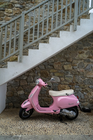 Mykonos, Greece - September 29, 2017: Beautiful classic design pink Vespa scooter with white seat cushion parking on gravel wash sidewalk under white and grey stair with stone wall background, Mykonos islandのeditorial素材