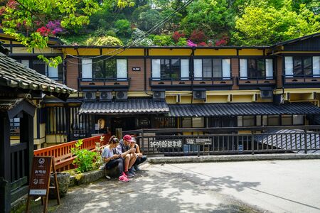 Kyushu, Japan - May 11, 2017 : Tourists sitting on plant pot on street with rail, local buildings and view of colorful blooming flowers and green trees upon hill background, Kurokawa onsen town, Kyushuのeditorial素材