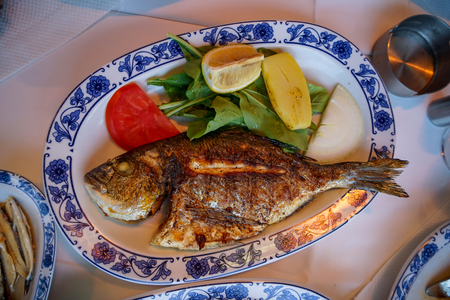 Fresh grilled whole sea bream fish with roasted potato, green salad, tomato and yellow lime on white oval plate with blue pattern at Galata bridge, Istanbul, Turkeyの写真素材