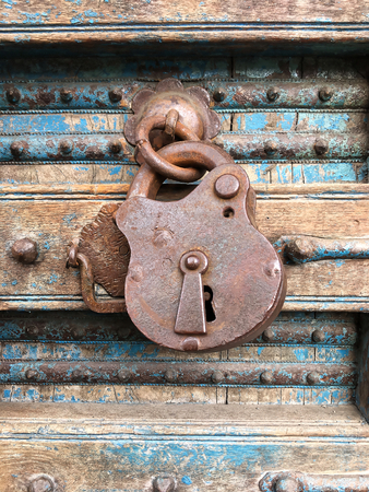 Old rusty padlock in the closed position, hanging on a dilapidated wooden door, with iron ornaments and peeling blue paintの写真素材