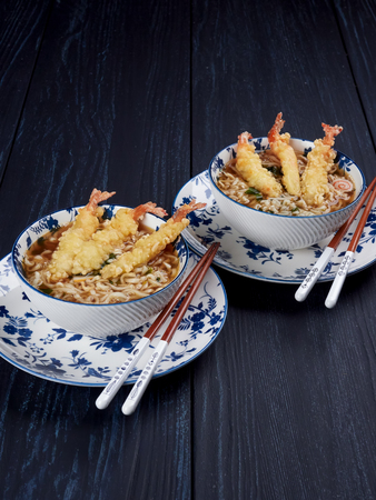 Two bowls of tempura udong noodles, with shrimps tempura, in white and blue bowls and chopsticks, on dark blue backgroundの写真素材