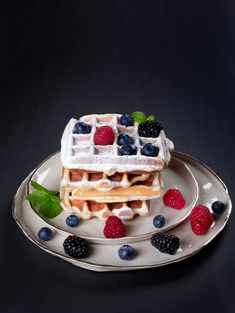 A stack of four home made Belgian waffles dusted with sugar, with fresh berries and mint leaves, on a beige platter, set on dark brown backgroundの写真素材