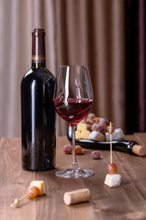 A bottle of red wine, a glass with red wine, a board with snacks, cheese and grapes on a wooden...
