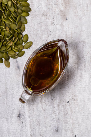 Glass gravy boat with pumpkin oil and pumpkin seeds on a gray concrete background, top view, close-up, selective focus on the oilの写真素材