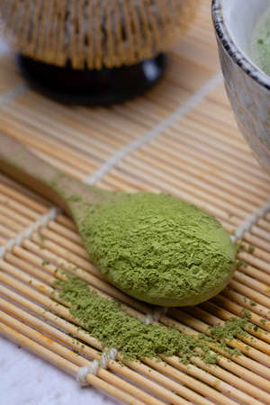 Traditional Japanese green tea match close-up. Focus on dry matcha tea in a wooden spoon, whisk for whipping and tea in a cup in the background in blur, a wooden background and a bamboo mat. Japanese style.の写真素材