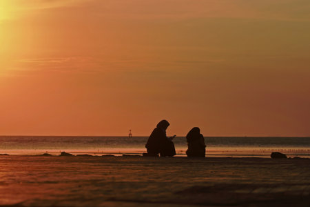 Two Muslim women captured in silhouette while watching a peaceful sunrise at the beach in Phuket, Thailand, with warm golden light reflecting across the calm shoreline.の写真素材