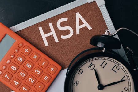 Top view of clock, calculator and brown board written with text HSA stands for Health Savings Account.の写真素材