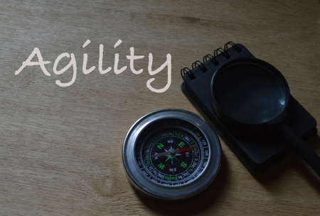 Top angle view of notebook, magnetic compass and magnifying glass over wooden background written with text AGILITY.の写真素材