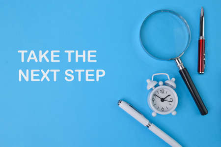 Phrase TAKE THE NEXT STEP written on blue background with pens, clock and magnifying glass.の写真素材