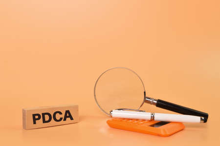 Magnifying glass, pen, calculator and wooden block written with PDCA stands for Plan, Do, Check,...