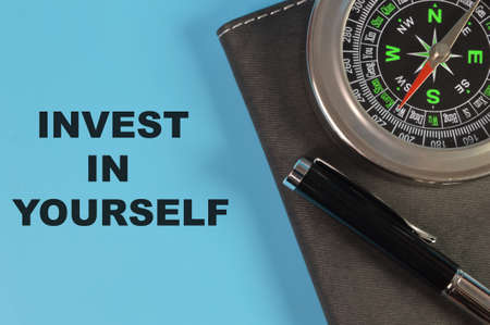 Compass, pen and notebook on blue background written with INVEST IN YOURSELF.