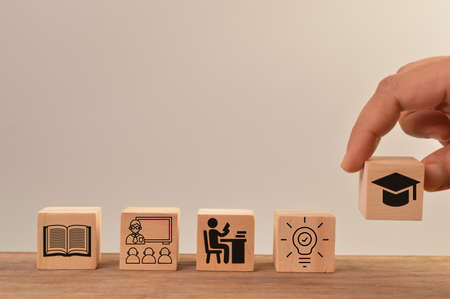 Hand holding graduation cap with bulb, student, classroom and book icons on wooden blocks. Key to...