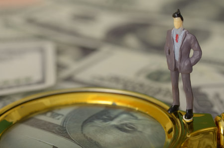 a miniature businessman standing on a magnifying glass, surveying a pile of dollar bills. The businessman seems to be searching for something specific, perhaps an opportunity or a challenge.の写真素材