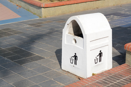 A white, arched trash can is placed on a paved walkway in a public park.の写真素材