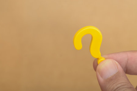 Close-Up of Hand Holding Yellow Question Mark on Tan Background â Concept of Inquiry and Curiosityの写真素材