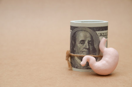 A human stomach leans against a roll of one-hundred-dollar bills, powerfully illustrating the grim...