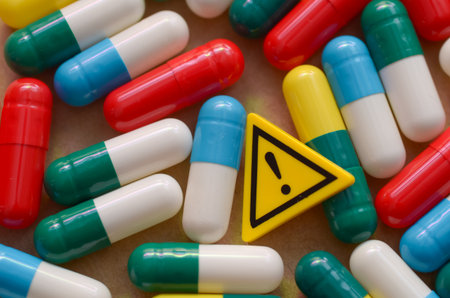 A pile of colorful medication capsules next to a caution sign with an exclamation mark, symbolizing the potential dangers and side effects of drugs.の写真素材