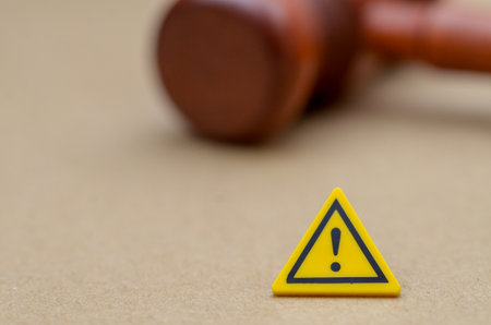 A sharp yellow warning sign in the foreground with a blurred judge's gavel behind it, symbolizing...