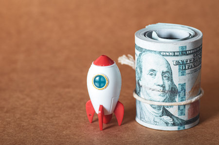 A rocket poised for launch next to a substantial roll of one-hundred-dollar bills, symbolizing the immense financial investment required for high-growth ventures and innovation.の写真素材