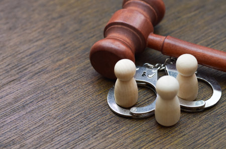 Three wooden figures stand within handcuffs before a judge's gavel, symbolizing a family or team facing a shared legal crisis and the consequences of a collective judgment.の写真素材