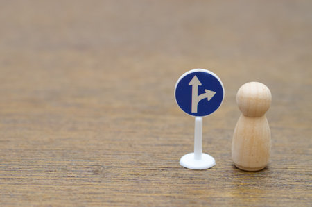 A wooden peg doll stands at a road sign with straight and right turn options, symbolizing a critical life or business decision about the future path.の写真素材