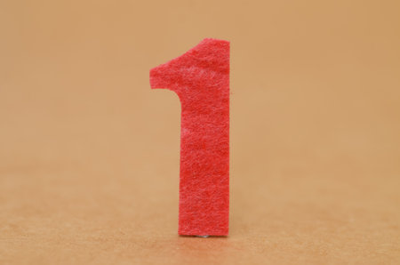 Red Felt Number One: A bright red, textured felt number "1" stands on a smooth, tan surface, symbolizing a start, a win, or the first place.の写真素材