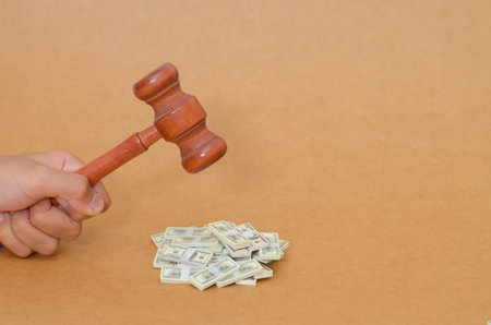 The Price of Justice: Firmly Grasped Gavel Poised Mid-Swing Over Miniature Stacks of Cash, Symbolizing Fines, Settlements, and the Intersection of Law and Finance.の写真素材