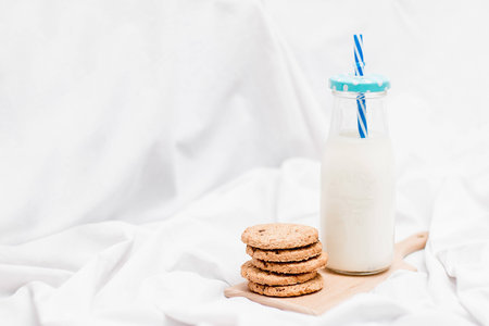 Photo of a jar of milk with blue straw and homemade cookies on a wooden board with white backgroundの写真素材