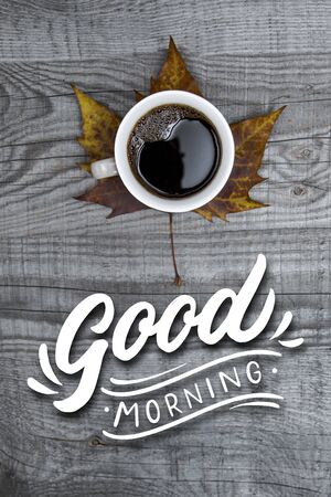Top view of a black coffee standing on a big autumn leaf on a wooden table with text Good Morning