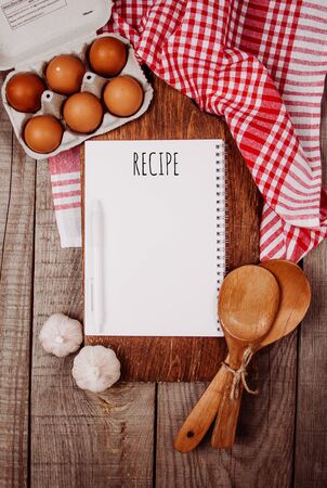 Wooden cutting board on a wooden background with garlic, ladles, eggs and a blank notebook with quote Recipeの写真素材