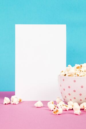 Fresh popcorn in a white pink bowl on pink and blue background with the blank paper pageの写真素材