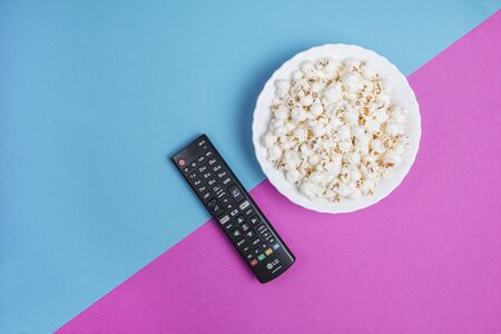 White bowl of popcorn on a blue and purple background with a black TV remote controlの写真素材