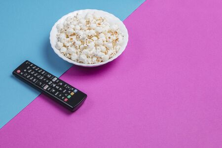 White bowl of popcorn on a blue and purple background with a black TV remote control