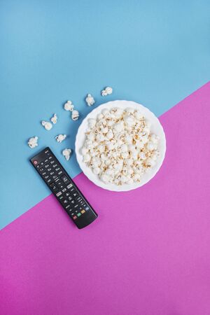 White bowl of popcorn on a blue and purple background with a black TV remote controlの写真素材