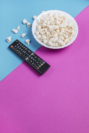 White bowl of popcorn on a blue and purple background with a black TV remote controlの写真素材