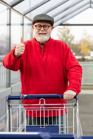 Smiling senior with a cap and red jacket pushing a shopping cart and make thumbs upの写真素材