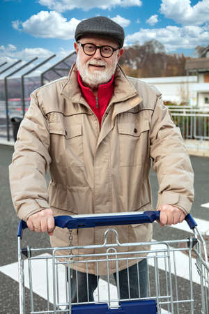 Smiling senior with a cap and beige jacket pushing a shopping cartの写真素材