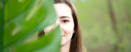 Beautiful natural young woman closeup behind big monstera leaf with green background in the woods. Beauty, health and nature concept.の写真素材
