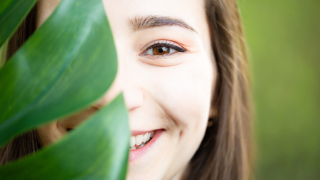 Beautiful natural young woman closeup behind big monstera leaf with green background in the woods. Beauty, health and nature concept.の写真素材