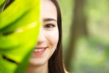 Beautiful natural young woman closeup behind big monstera leaf with green background in the woods. Beauty, health and nature concept.の写真素材