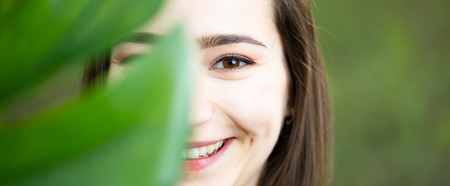 Beautiful natural young woman closeup behind big monstera leaf with green background in the woods. Beauty, health and nature concept.の写真素材