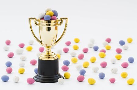 A gold trophy full of small, chocolate Easter eggs for the winner of an Easter egg hunt. Small candy Easter eggs scattered over an isolated white background.の写真素材